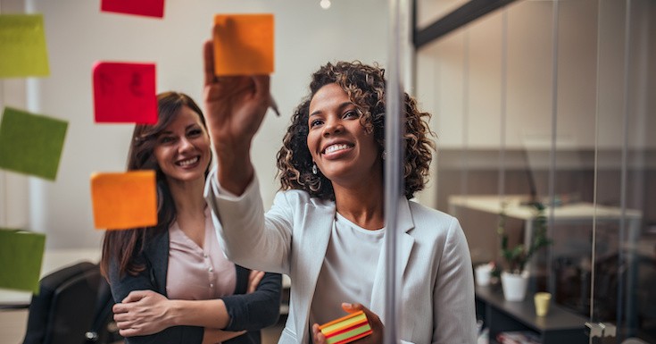 Creative businesswoman writing on sticky notes on a glass wall, female colleague looking.